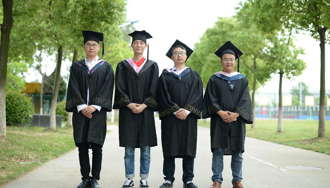 Group of four graduates wearing caps and gowns standing outdoors on a bright day.
