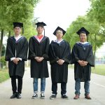 Group of four graduates wearing caps and gowns standing outdoors on a bright day.