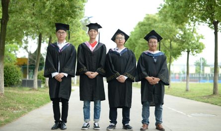 Group of four graduates wearing caps and gowns standing outdoors on a bright day.