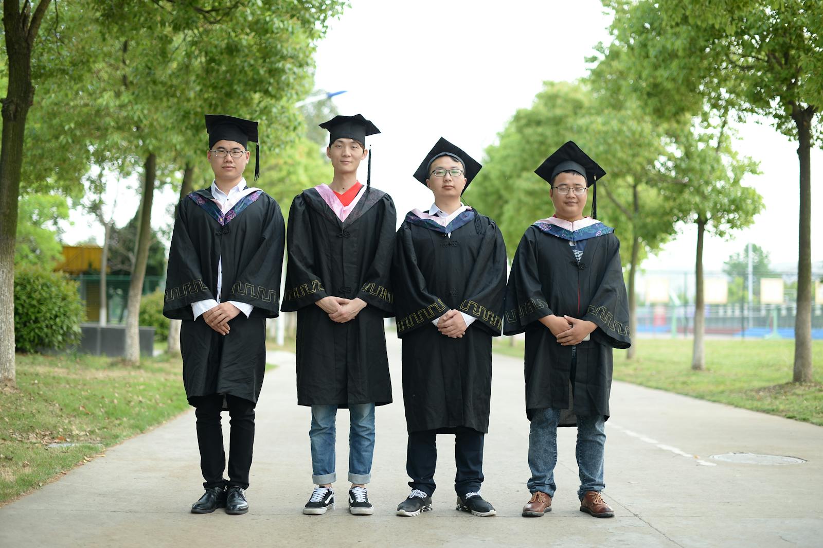 Group of four graduates wearing caps and gowns standing outdoors on a bright day.