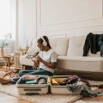 Young woman sits at home packing a suitcase while listening to music, surrounded by clothing items.