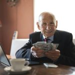 Elderly man smiling while counting cash at a home office setting.
